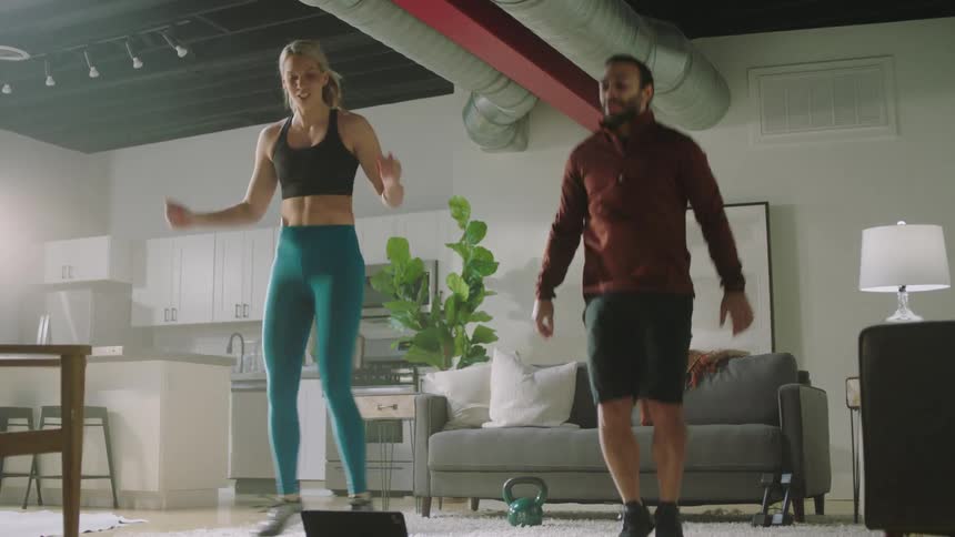 Blonde woman with black sports bra and teal leggings works out next to man with beard and shirt in living room at home