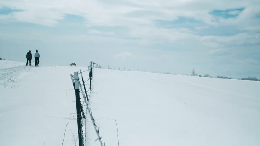 Man and woman wearing snowshoes walk on a snowy hill near a fence with a dog under a cloudy blue sky