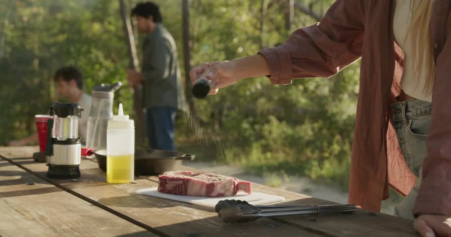 Woman sprinkles seasoning on a steak that is resting on a picnic table while two men are nearby on a sunny day