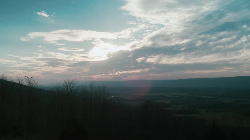 Trees grow on a hill with a valley below and mountains in the distance under a cloudy sky