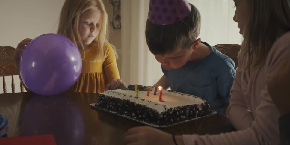 A woman carries a cake with candles and places in front of a little boy at a table who blows them out