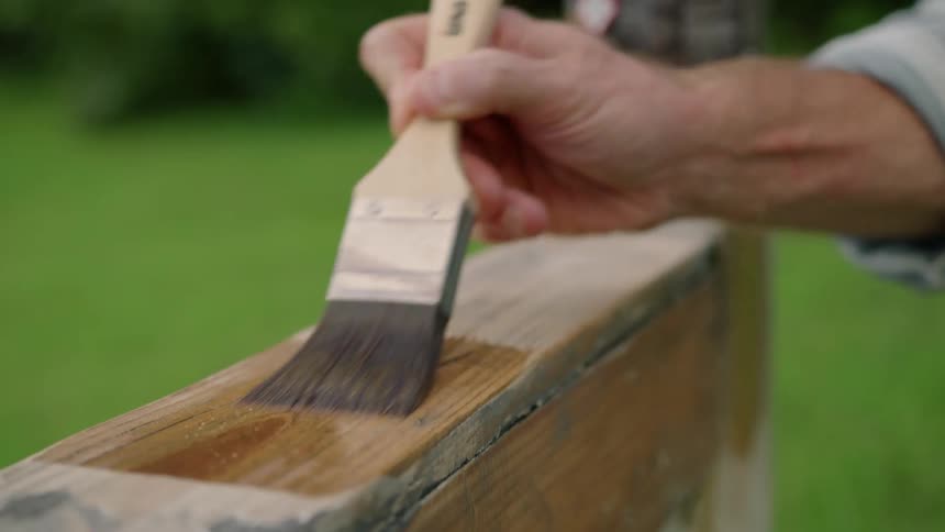 A hand stains a wooden object with a paintbrush outdoors