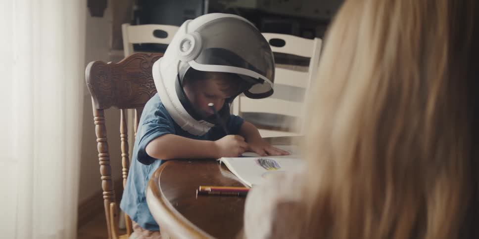 A young boy coloring at a dining table while wearing a space helmet with his mom