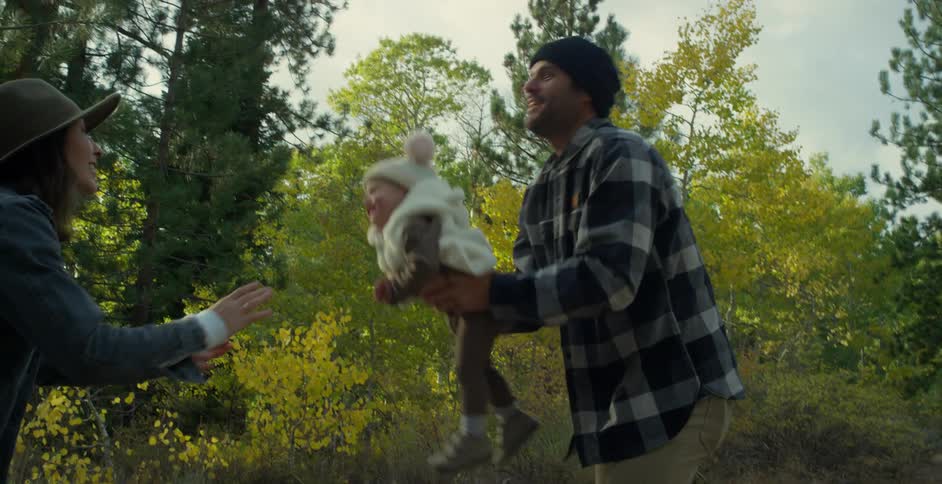 Mother with brown hat wavy hair and denim jacketand father wearing green wooly hat and gray plaid shirt lift toddler baby up in in autumn forest