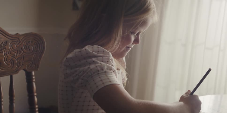 Blonde girl wearing white dress with red dots and sitting at table draws in drawing book