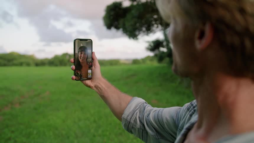 Blonde man with gray shirt makes video call with woman blowing kiss at each other on green farm