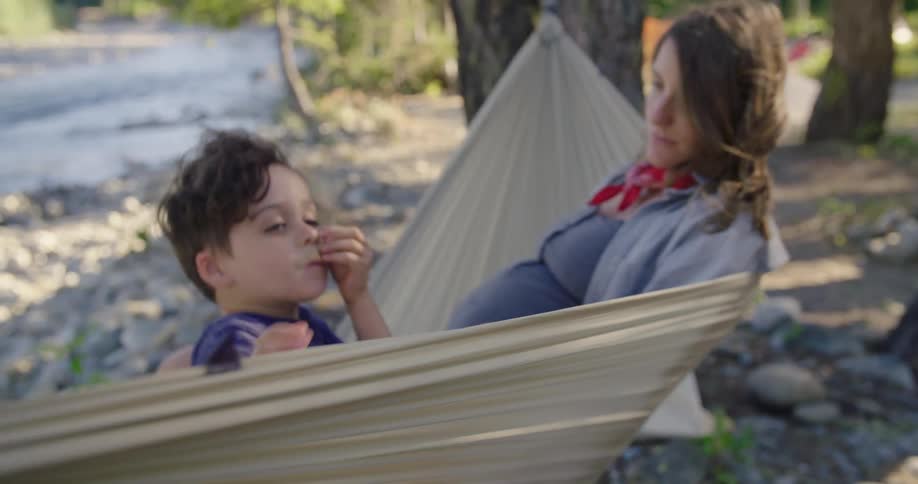 Pregnant mother with wavy hair and son sit in hammock on river bank