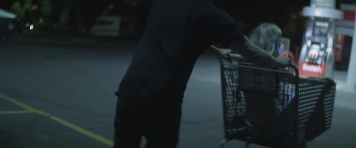 A man pushes a girl around a gas station parking lot in a shopping cart