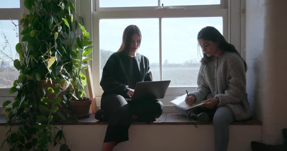 Two women are sitting at a window working