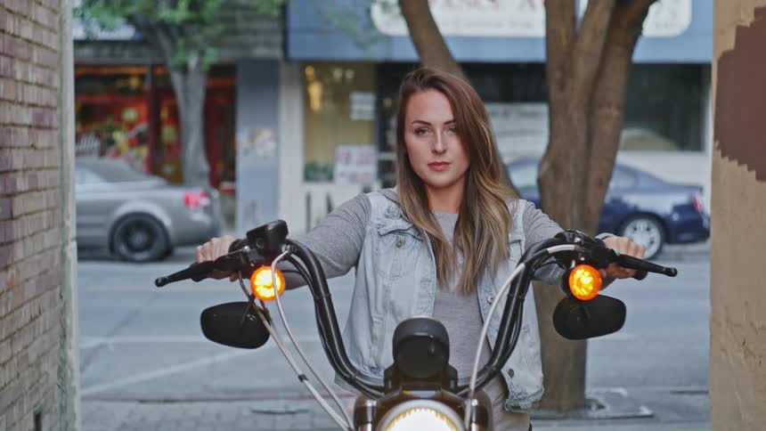 A young woman with long hair sits atop a parked motorcycle