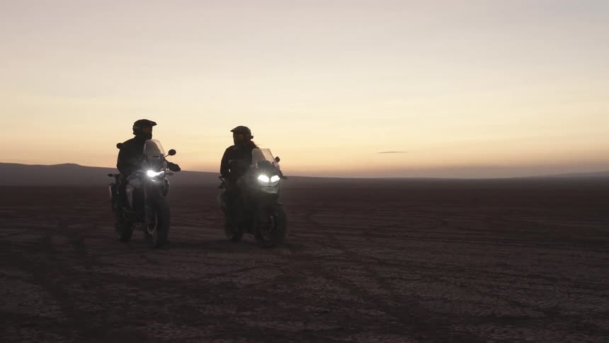 Two motorcyclists sit on motorcycles next to each other in the desert at dawn