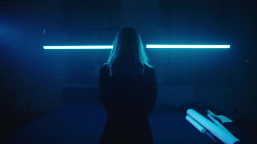 Blonde woman walks toward table and spreads rolled paper onto it under a fluorescent light