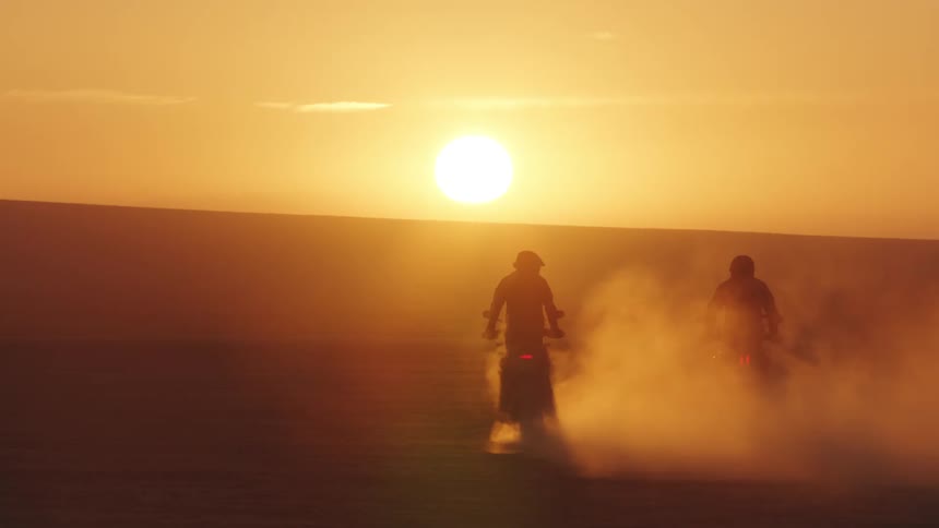 Two people drive over a dusty landscape on motorcycles while the sun is low in the sky