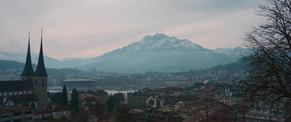 Lake and buildings in a city sit near the snow covered mountains under a cloudy sky