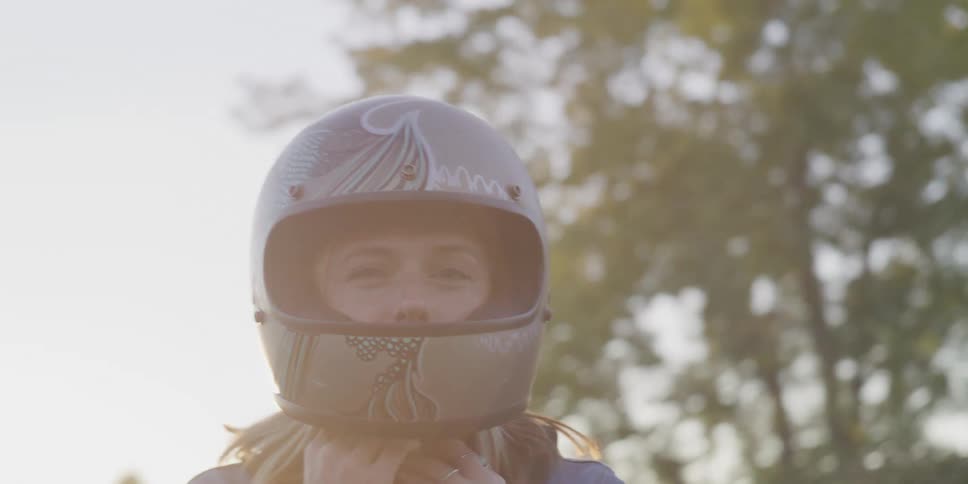 Blonde woman with silver motorcycle helmet faces the camera with eye contact in front of trees