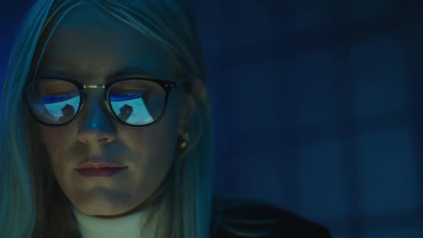 Young blonde woman wearing glasses drawing on a touchscreen with a stylus on a dimly lit room
