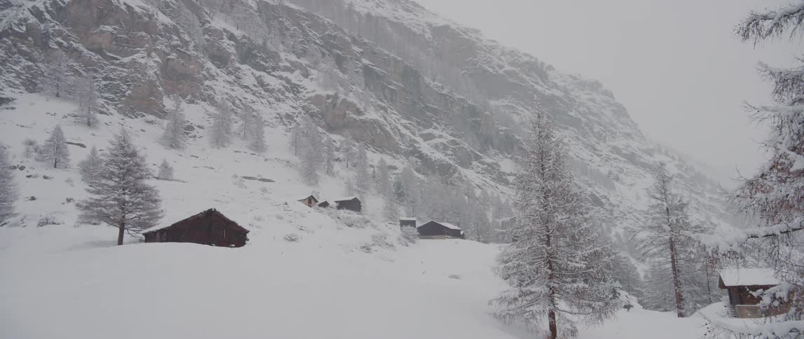 Snow falling over cabins scattered at the bottom of a tall mountain at winter