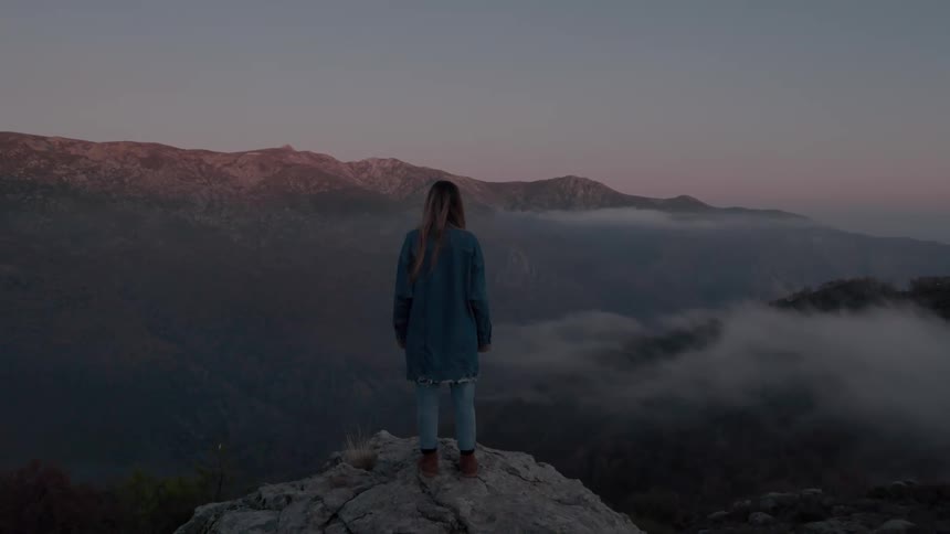 A woman stands perched on a mountain rock overlooking a forest in a valley and low clouds at dawn