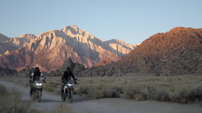 Two motorcycles driving on a dirt road in the desert close to the mountains at evening