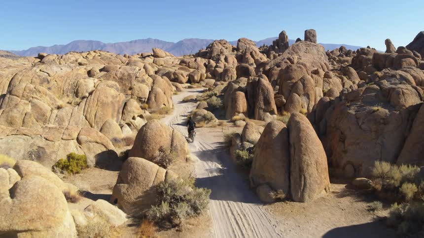 A person drives a motorcycle down a dirt road past large rock formations