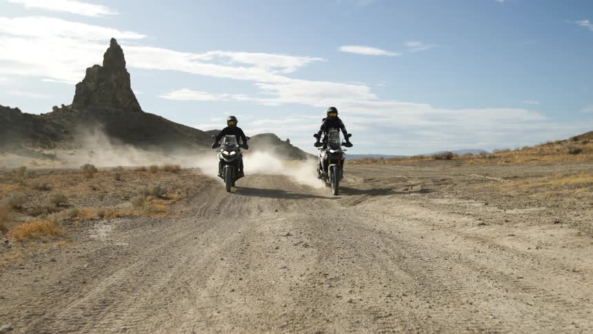 Two motorcyclists ride motorcycles on a sunny day on a dirt road that runs through a field