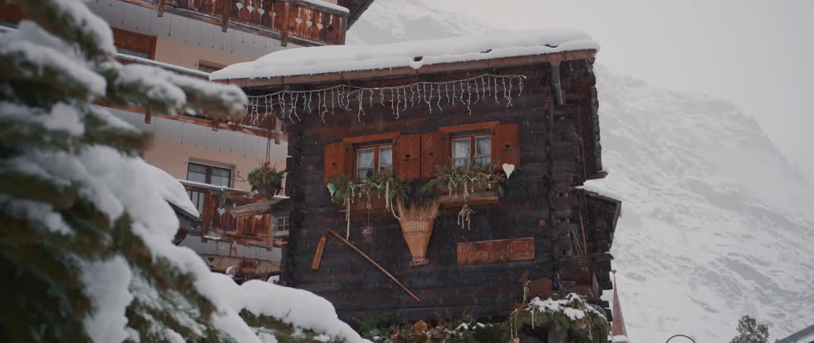 Snow falling over a small cabin next to a building close to the mountains at winter