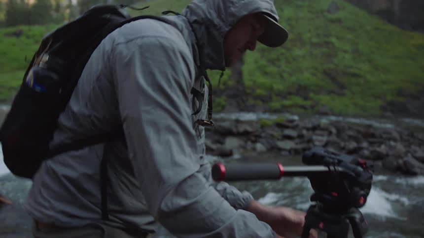 Male photographer with gray raincoat black backpack and brown pants installs camera equipment next to river bank