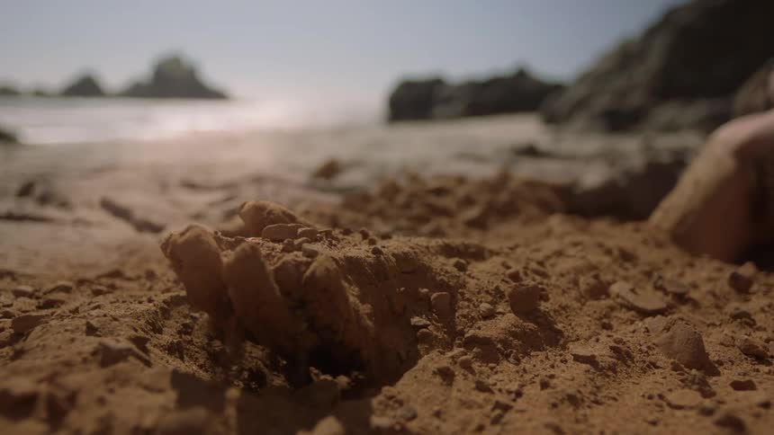 A man with his arm buried under the sand on a beach slowly raises his arm to release it from the sand