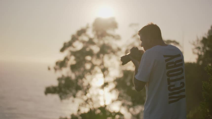 Man wearing a short sleeved shirt takes a photo of a lake using a camera with a long lens attachment on a sunny day