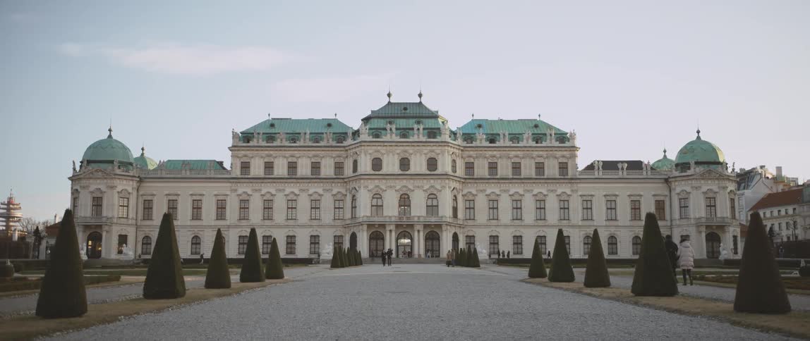 Wide landmark with many windows with two rows of triangular shaped trees on each side of the pathway in front