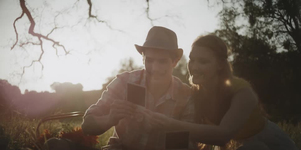 A man and woman look at a polaroid photo as they sit outside in the grass