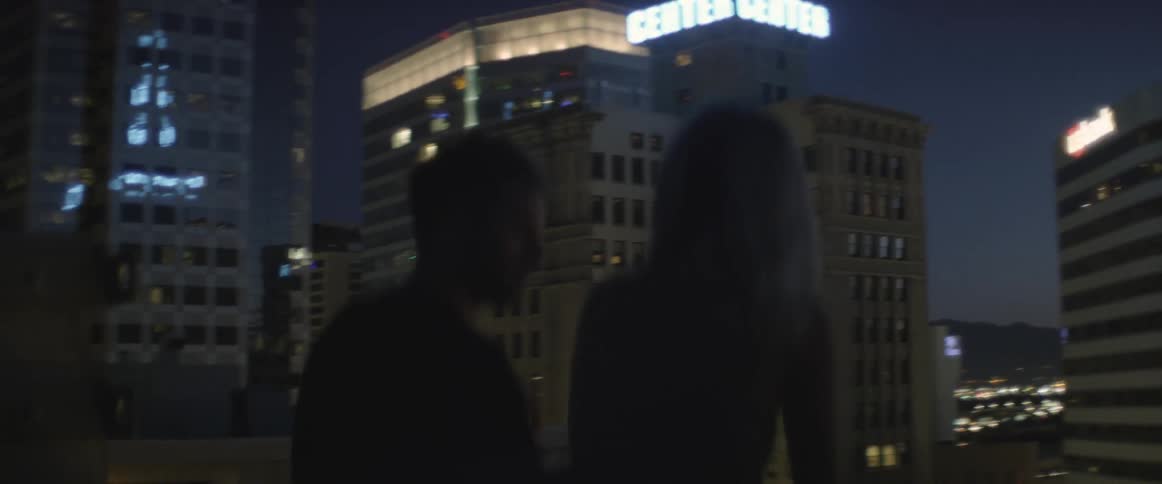 A man and woman talking on a rooftop in a city at night