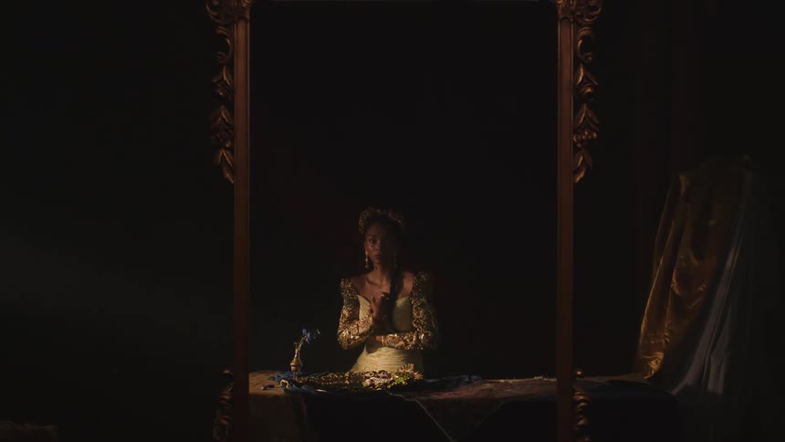 Woman in theatrical costume sits at table near vase with flowers framed by ornate wooden frame