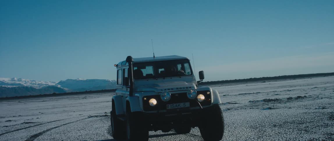 A land rover travels in a barren landscape with a snow covered mountain behind