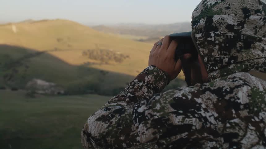 A man stands on a hill and looks through binoculars