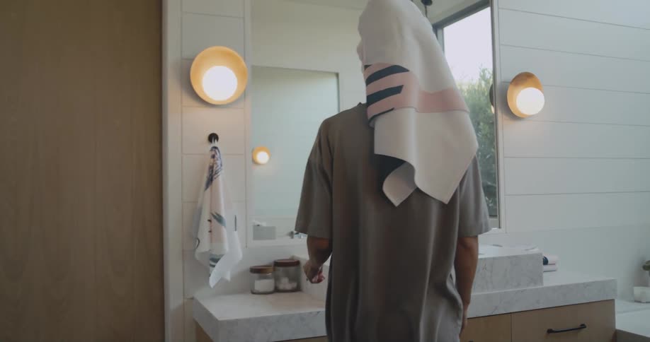 Woman with hair towel and gray shirt walks to kitchen sink