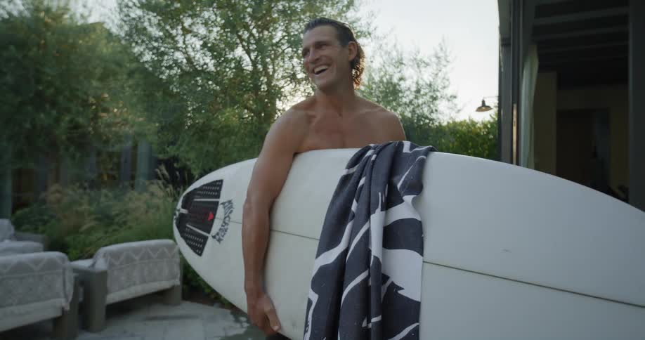 Shirtless man holds white surfboard and blue towel in front of building laughing in the garden