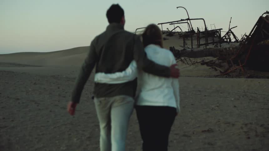 A couple walk with their arms around each other toward a desert shipwreck