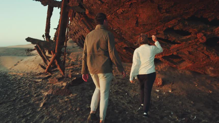 Man and woman walk towards a desert shipwreck and reach up to touch it