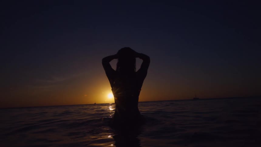 A woman in the ocean as the sun sets stands up and runs her hands through her hair