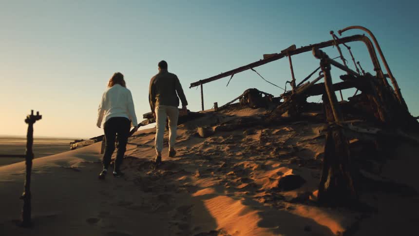 Man and woman walk up to a desert shipwreck at sunset