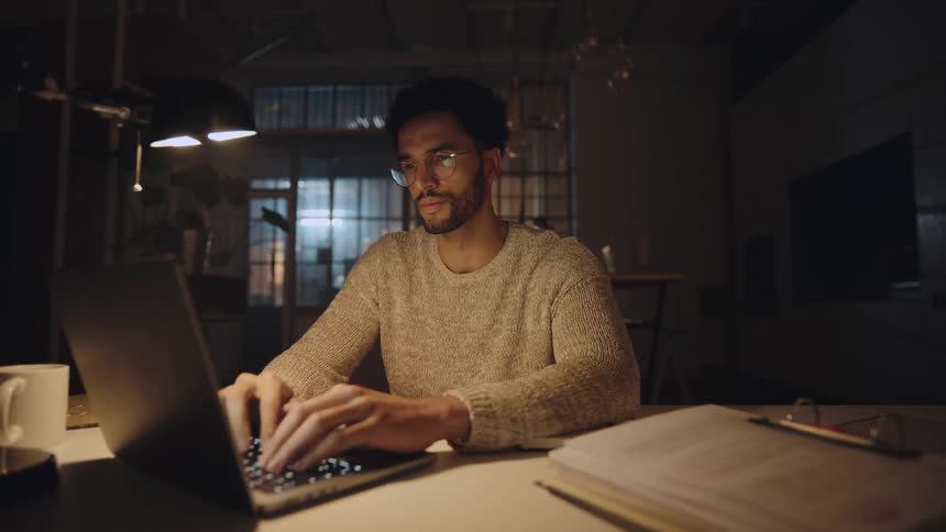 Man sits in office at night working on laptop while trying to ignore exhaustion
