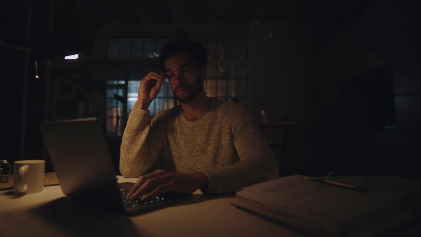 A man rubs his eye tiredly then continues to work on his laptop at night