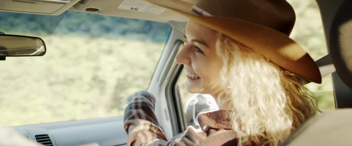 A woman in a cowboy hat laughs while riding in the passenger seat of a moving vehicle
