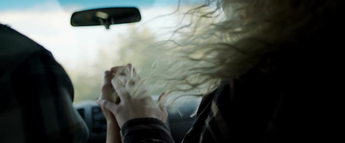 Man driving a vehicle holds hands with a blonde woman as they travel past the trees