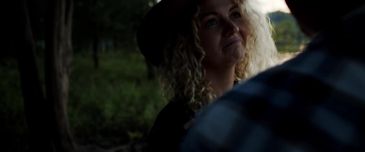 Woman wearing a brimmed hat hugs a man while they stand in the forest and look out at the lake