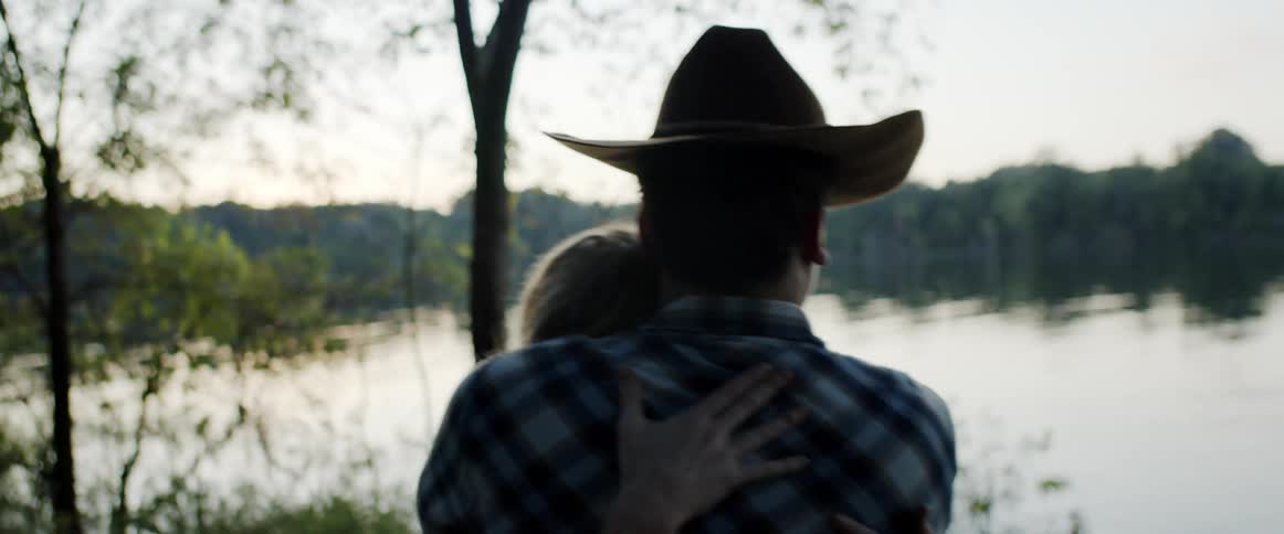 Man wearing cowboy hat hugs his partner near a lake in the forest at morning