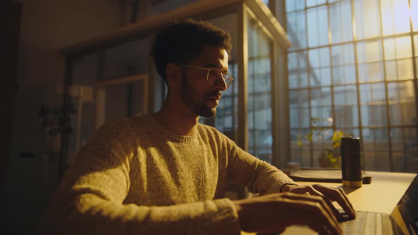 Young man sitting at a desk opens a laptop and starts typing in front of a window with sunlight coming in