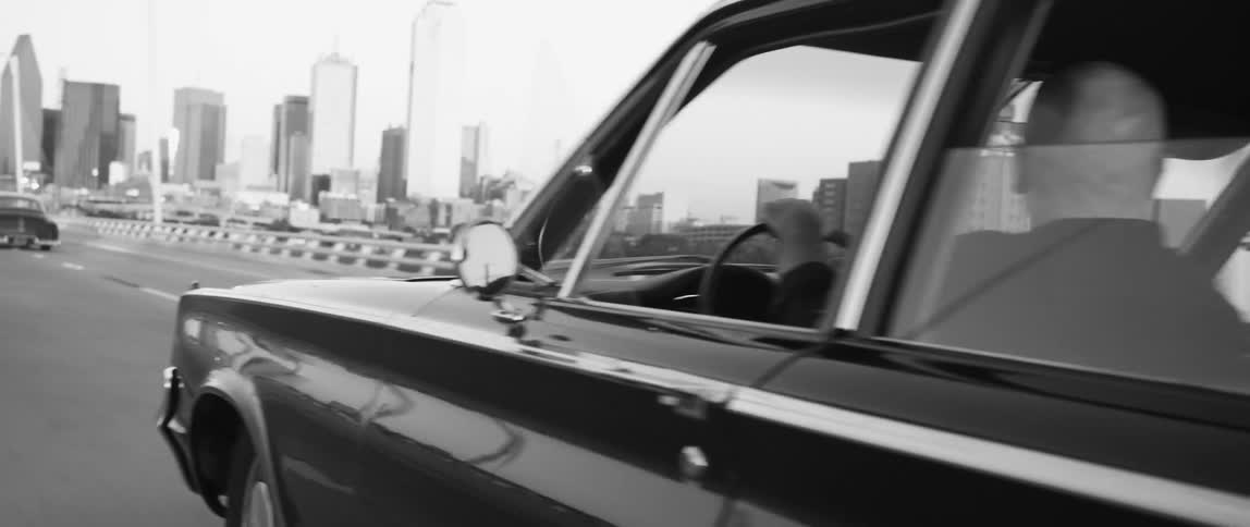 Black and white footage of vintage vehicles driving over a bridge near the city skyline at morning