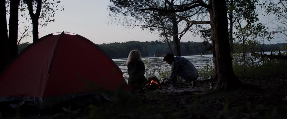 A man uses his hat to fan a campfire while camping beside a river with a woman